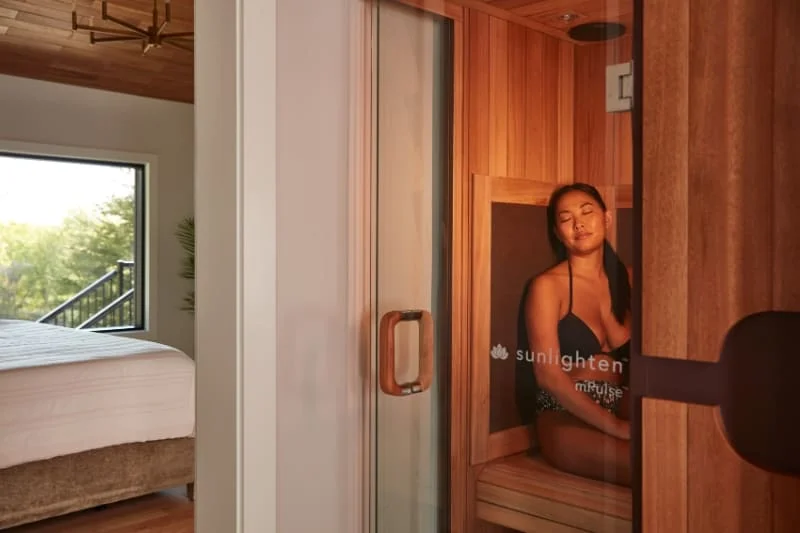 Woman enjoying a Sunlighten mPulse Believe infrared sauna installed in a bedroom setting, with glass door partially open showing the wooden interior, sauna controls, and comfortable bedroom furnishings visible in background.