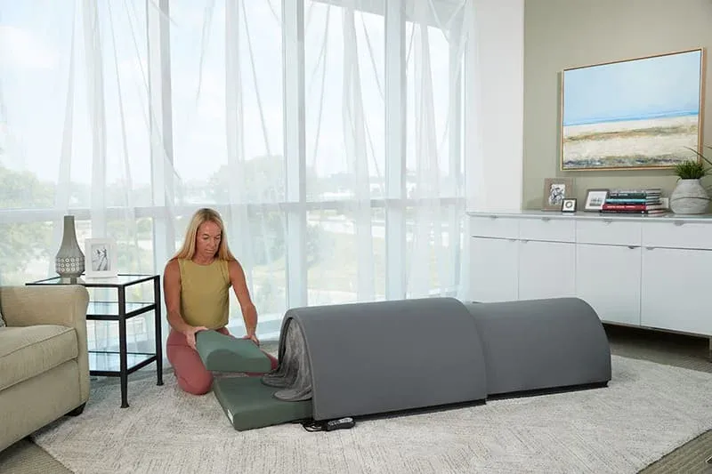Woman assembling the Sunlighten Solo portable infrared sauna in a bright living room with large windows.
