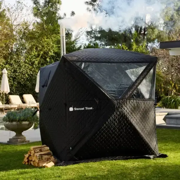 A black quilted Sweat Tent portable sauna set up on a grassy backyard with steam coming from the chimney. There's a stack of firewood nearby and lounge chairs visible in the background.