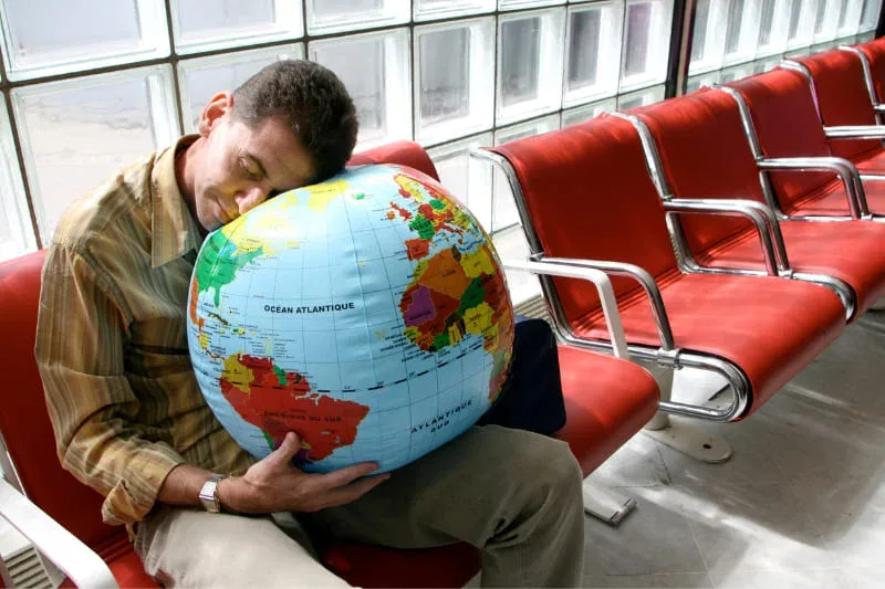 Exhausted traveler resting head on globe in airport waiting area, showing physical fatigue from international travel.