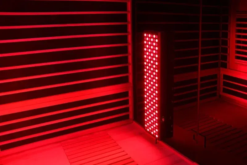 Full-length vertical red light therapy panel mounted between wooden slat walls in a sauna, illuminating the bench and floor area.