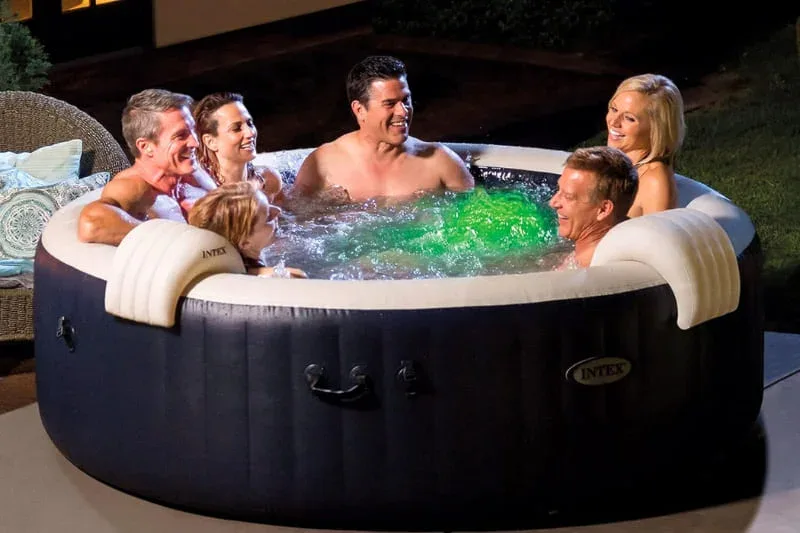 Group of adults enjoying Intex inflatable hot tub at night with LED lighting, navy blue portable spa with green illuminated water and outdoor patio furniture