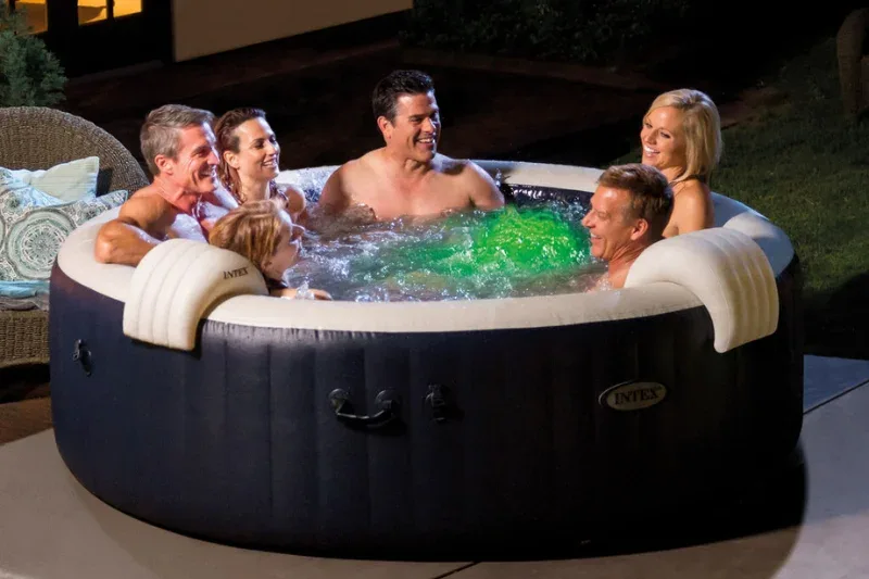 Six adults enjoying Intex inflatable hot tub with green LED lighting on outdoor patio at night