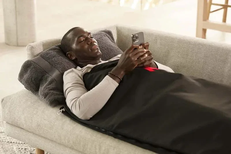 Man relaxing in MiHigh infrared sauna blanket on couch while using smartphone during home wellness session