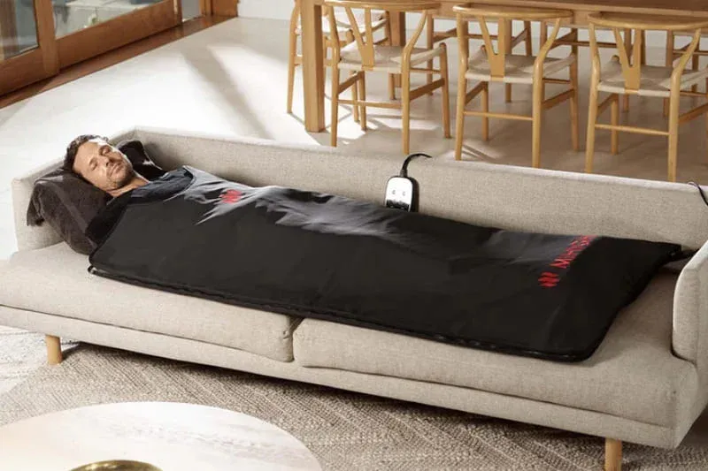 Man lying in MiHigh infrared sauna blanket on living room couch with handheld controller for at-home heat therapy