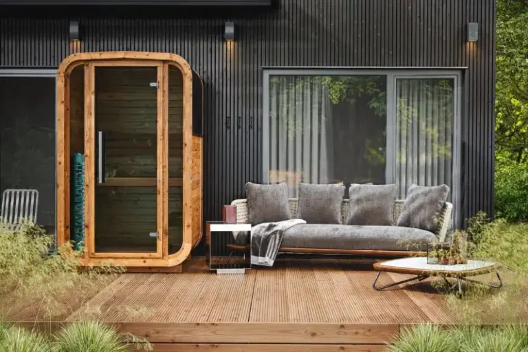 A traditional wooden barrel sauna from Auroom with glass door sits on a wooden deck beside a modern gray building. The curved cedar sauna features a glass front panel, and the deck area includes a gray outdoor sofa with cushions and a low coffee table, surrounded by natural landscaping.