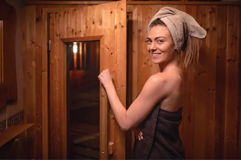 Smiling woman in towel entering wooden sauna door, showing proper nude sauna etiquette