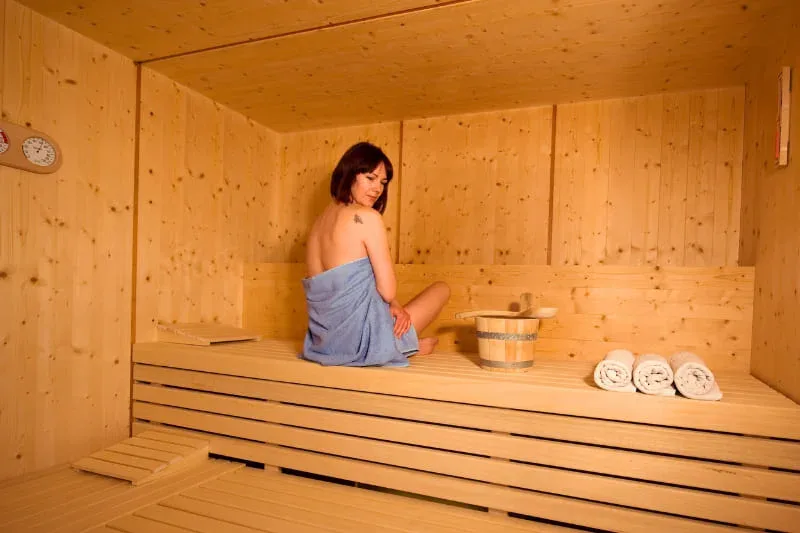 Woman sitting on towel in wooden sauna with traditional bucket and rolled towels nearby