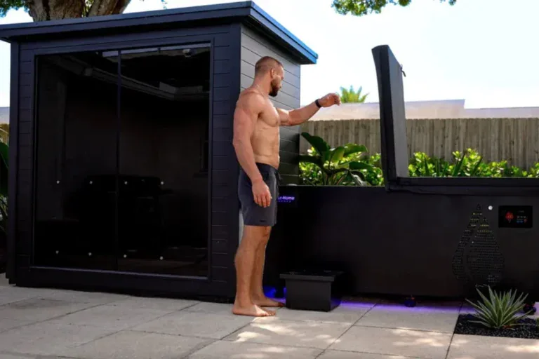 Man standing between an outdoor sauna and a cold plunge tub, showing a modern home wellness setup for contrast therapy.